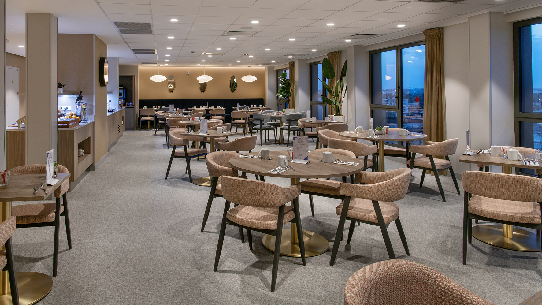 Salle de cantine avec des tables rondes et des chaises, fenêtres et bar au fond de la salle